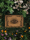 A 24x16 inch rectangular outdoor doormat made of eco-friendly coconut coir with a black vinyl backing. The mat features a central evil eye design radiating with detailed rays and surrounded by swirling ornamental scrollwork and small stars. Designed for witchy home entryways, it offers protection and style for porches, patios, and thresholds.
